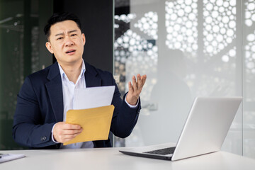 Annoyed asian businessman looking at camera and showing received letter with bad news, man working inside modern office building, using laptop.