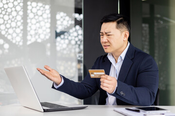 Frustrated asian man trying to make banking transaction and purchase in online store, businessman cheated and sad working inside office with laptop at work holding bank credit card.