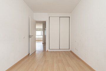    An empty room in an apartment with a built-in wardrobe with white sliding doors, a light wooden floor and white access doors