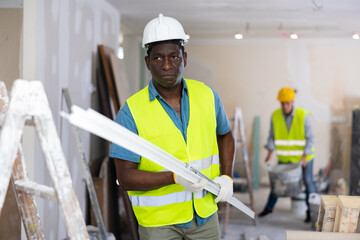African-american repairman wearing protective hardhat and yellow vest carrying construction detail...