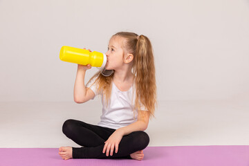 Beautiful little child girl in white t-shirt, sitting on fitness mat with a yellow sports bottle of water in her hands. For body hydration after workout indoors or yoga practice at home