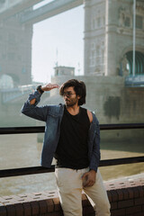 Indian man standing on Tower Bridge. He wears a denim jacket, sunglasses and he is looking away from camera.