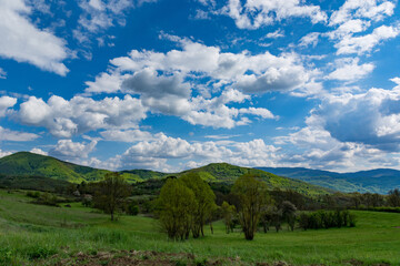A beautiful natural landscape featuring green hills bathed in sunlight and lush meadows, with beautiful white fluffy clouds dotting the sky on a spring day