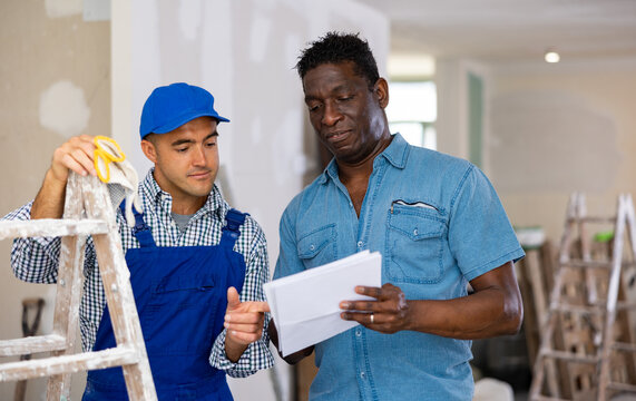 African-american man owner of apartment and caucasian man builder discussing project documentation in apartment during repair works.