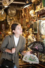 Young woman tourist in casual clothes chooses vintage fan in antique shop