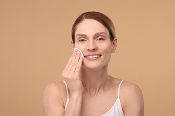 Beautiful woman removing makeup with cotton pad on beige background