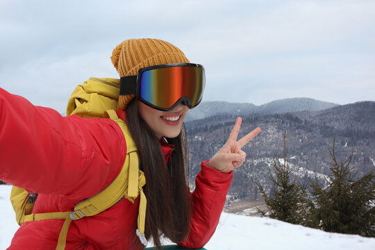 Smiling woman in ski goggles taking selfie in snowy mountains