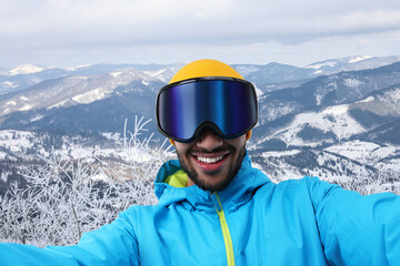 Fototapeta na wymiar Smiling young man in ski goggles taking selfie in snowy mountains