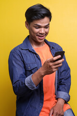 Excited adult Asian man looking at mobile phone and laughing on yellow background