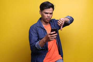 Asian man give bad rating and showing disappointed expression while holding mobile phone isolated on yellow background