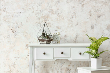 Florarium with alarm clock and candle on table near light wall