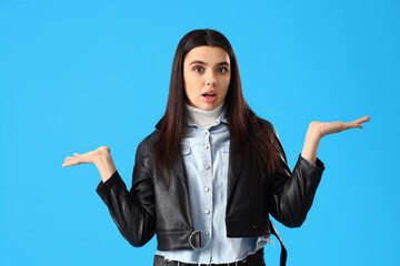 Confused young woman in leather jacket on blue background