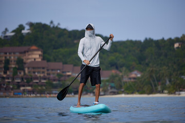 Guy in white hoodie and black shorts standing on board with paddle in hands, floating withf tropical coast on background, searching for wave, happy holiday concept, copy space