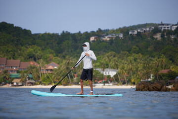 Surfer in white hoodie and black shorts standing on board with paddle in hands, floating with tropical coast on background, sport life concept, copy space