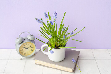 Cup with beautiful Muscari flowers, alarm clock and book on tiled table near lilac wall