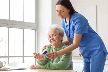 Obraz premium Young caregiver measuring temperature of senior woman with infrared thermometer in kitchen