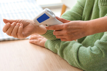 Young caregiver measuring temperature of senior woman with infrared thermometer in kitchen, closeup