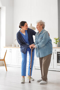 Young caregiver helping senior woman with walking stick in kitchen