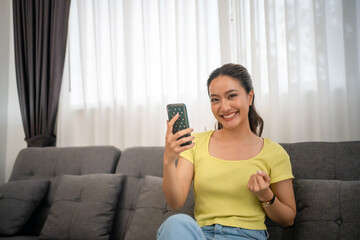 Asian woman sitting on sofa at home with mobile phone and feeling excited and happy.