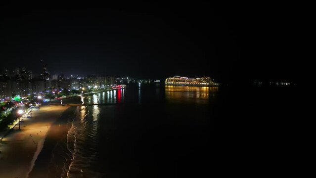 Illuminated cruise ship in the city at night. Coastal city illuminated at night. Aerial view of cruise ship leaving Santos city.
