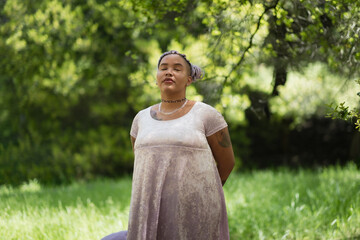 A strong nonbinary Afro-Latinx model poses with pride and assertiveness. Their purple braids and radiant makeup create an eye-catching contrast against the background of lush, leafy foliage.