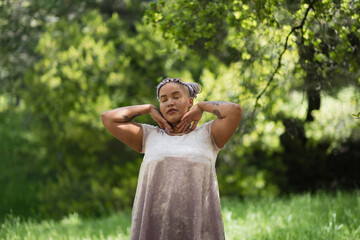 With their eyes closed, a nonbinary Afro-Latinx model with purple braids and a cute dress stands in a clearing. They have their hands by their cheeks, expressing a cute, lighthearted pose.