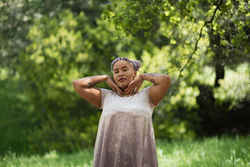 With a direct gaze, a nonbinary Afro-Latinx model with purple braids stands in a forest clearing. They are expressing a cute, lighthearted pose with their hands against their cheeks.