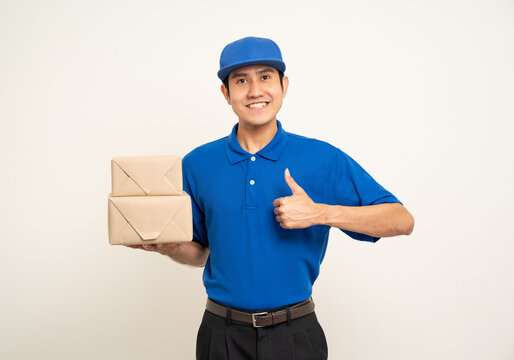 Happy delivery asian man in blue uniform standing holding box parcel cardboard on isolated white background. Smiling male service worker. Delivery courier and shipping service.