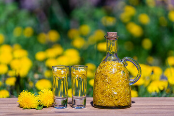 Homemade dandelion flowers tincture in two glasses and in a glass bottle on a wooden table in a summer garden, closeup