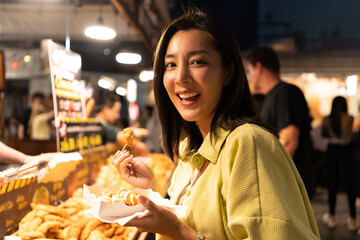 Naklejka premium Asian woman enjoy eating fries street food at night market. Traveler Asian blogger women Happy tourists Beautiful female with Traditional thailand bangkok food.