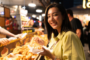 Obraz premium Asian woman enjoy eating fries street food at night market. Traveler Asian blogger women Happy tourists Beautiful female with Traditional thailand bangkok food.