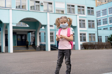 a little girl near the school with a backpack in a mask and an antiseptic in her hands, the concept of prevention covid-19