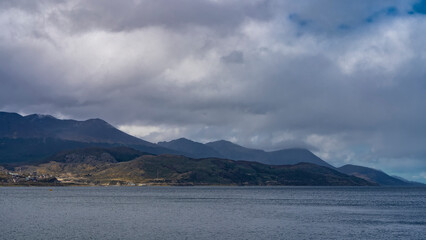 A picturesque mountain range against a cloudy sky. The blue water of the Beagle Canal in the foreground. City houses are visible on the shore. Argentina. Ushuaia. Tierra del Fuego archipelago.