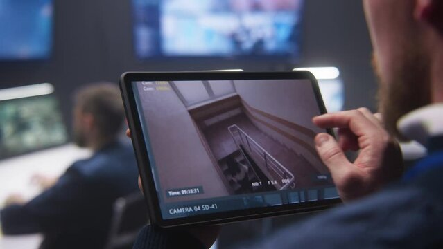 Close up shot of security officer zooming CCTV cameras with AI facial scanning using tablet in police surveillance center. Colleagues work at background. Monitoring system. Concept of social safety.