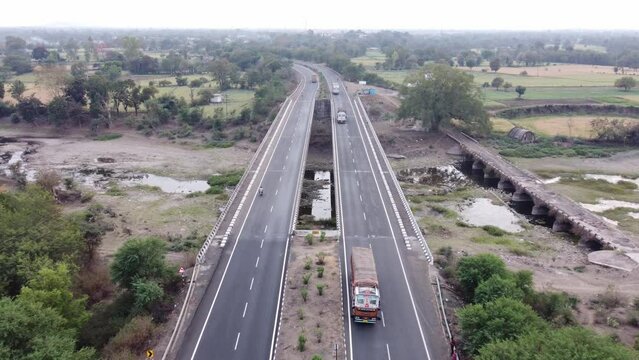 Drone coming down to establish a generic looking well maintained national highway of India with rural landscape of farmlands and river with heavy commercial goods carrier trucks driving by