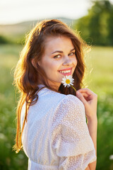 close portrait of a beautiful, laughing woman in a light dress holding a camomile near her face © VICHIZH