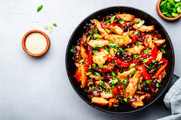 Asian cuisine, stir fry with chicken fillet, red paprika, mushrooms green onion and sesame in frying pan. Gray kitchen table background, top view, copy space