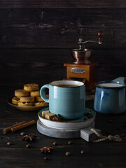 Blue clay cup with black coffee on a round wooden plank. Cinnamon tubes and star anise on the table. Biscuits and coffee grinder in the background. Dark wooden background