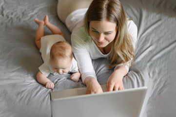 A young mother works remotely on a laptop, a 2.5-month-old baby lies on the bed next to her mother. Working mom.