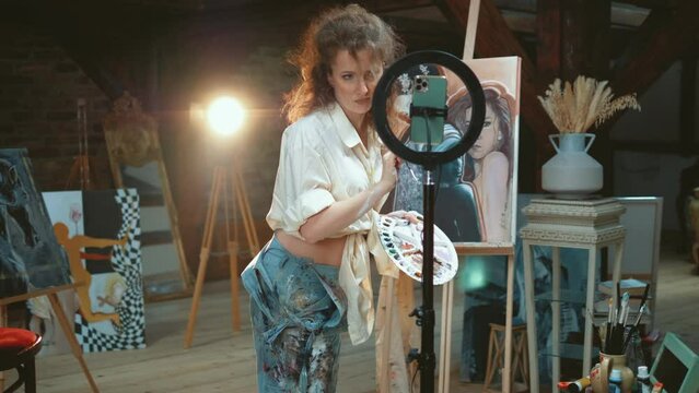 Beautiful art woman standing in the workshop next to the picture, reading comments on a phone while recording stream for vlog