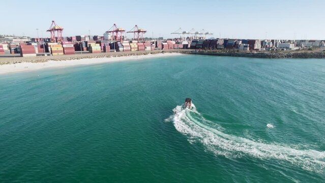 An aerial shot of a teenage boy Skurfing behind a speed boat at fremantle Port. The centre console boat is pulling the surfer behind the boat with the port visible in the back ground. 