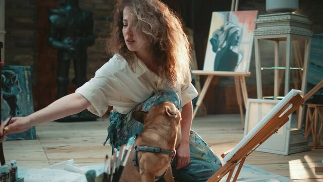 Slow motion shot of a dog kissing its owner, a woman painting on oil canvas in her art studio