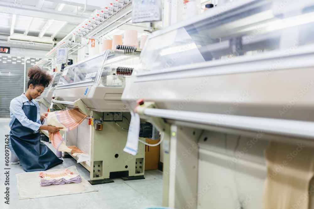 Wall mural american female designer wear work uniform sitting and inspecting the fabric in the weaving industry Around it there was a working sewing machine and multi-colored threads for fabric production.