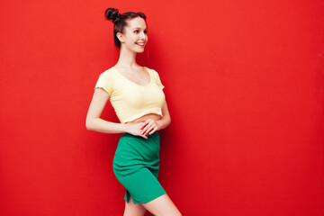 Young beautiful smiling female in trendy summer yellow t-shirt and green skirt. Carefree woman with two horns hairstyle posing near red wall in studio. Positive model having fun. Cheerful and happy