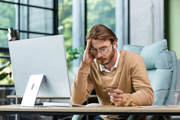 Cheated and disappointed businessman working inside office, man holding bank credit card and trying to make a purchase in online store, online fraud and deception