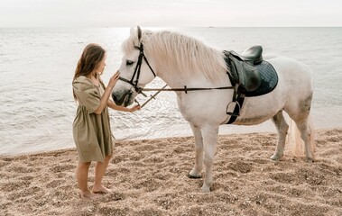 A woman in a dress stands next to a white horse on a beach, with the blue sky and sea in the...