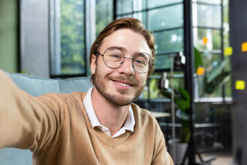 Portrait of successful businessman in casual shirt and glasses man looking at smartphone camera and smiling, camera view video call talking with friends inside office remotely