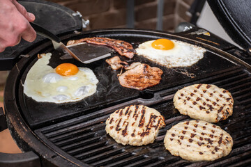 Burger patty, eggs and bacon grilling on barbecue grill plate . Backyard BBQ grill cooking burgers.