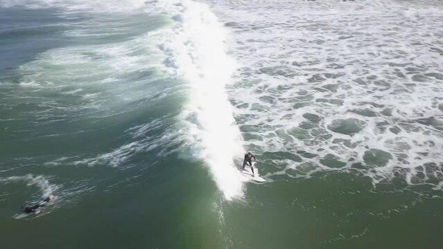 Man on a surfboard surfing as the drone flying with her on the wave on a very sunny day in cold water surf with an aerial view. Coxos surf spot.