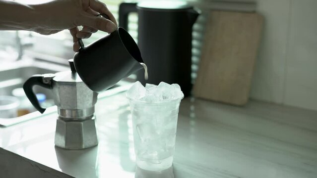 Pouring milk making latte iced coffee on plastic glass placed on a grey marble kitchen. preparation of coffee at home